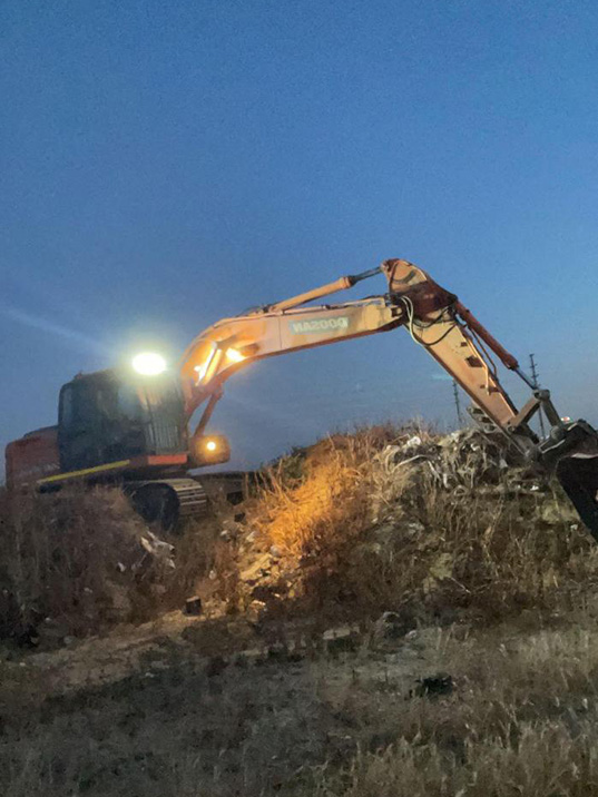 Excavator with its work lights on clearing scrub and debris at dusk