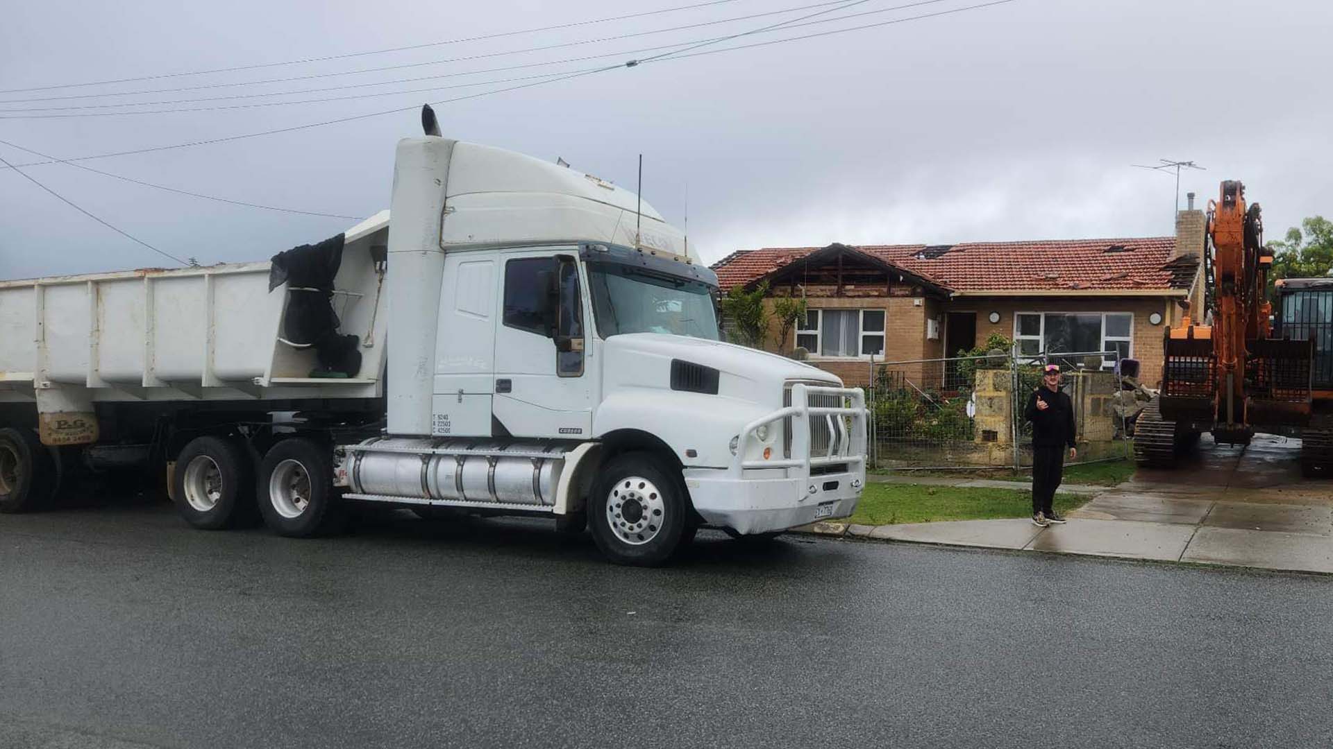 White tipper truck outside a brick house being demolished, excavator in the driveway